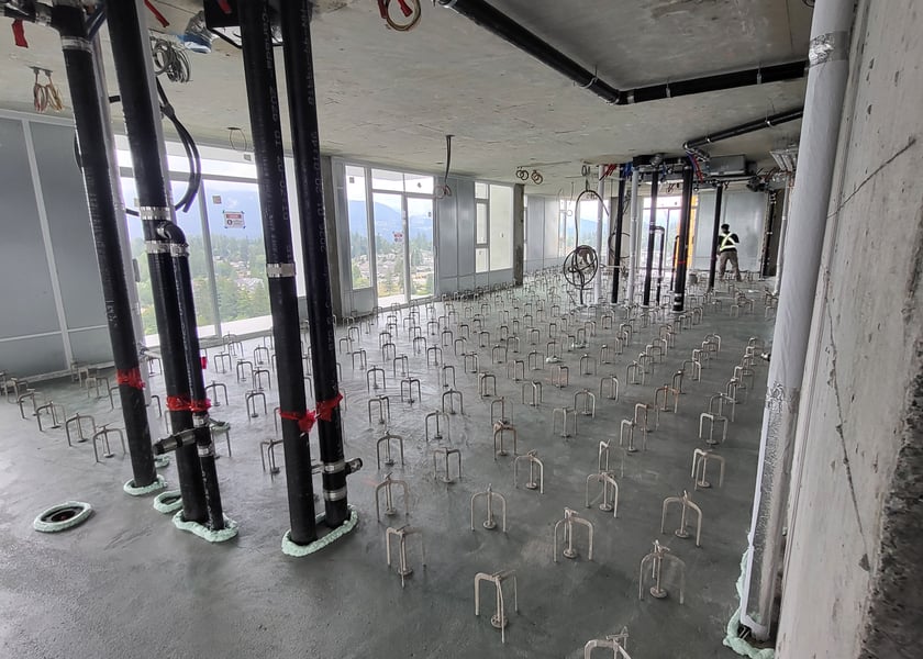 Rows of leveling pedestals across a concrete slab beside black pipe risers, mountain and city view through the window wall