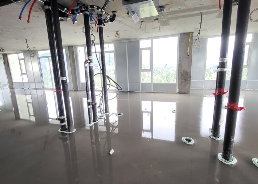 Self-leveling compound poured across a unit floor, reflecting the pipe risers and window wall