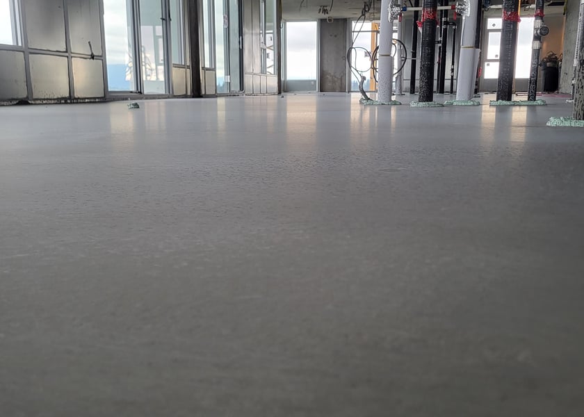 Low angle view of wet self-leveling compound on a concrete floor, pipe risers wrapped around columns in the background