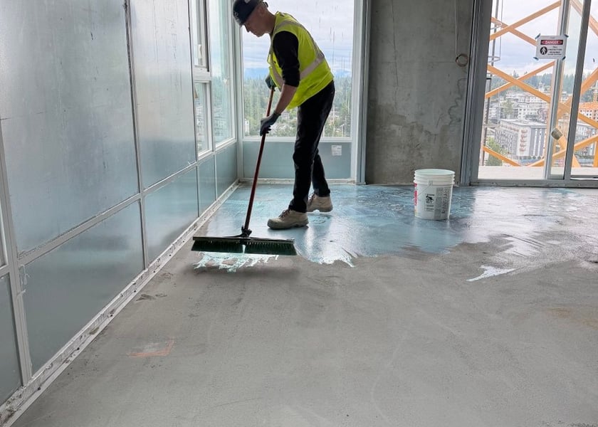Worker in a safety vest spreading blue tinted primer across a concrete floor with a push broom, a tower crane visible outside
