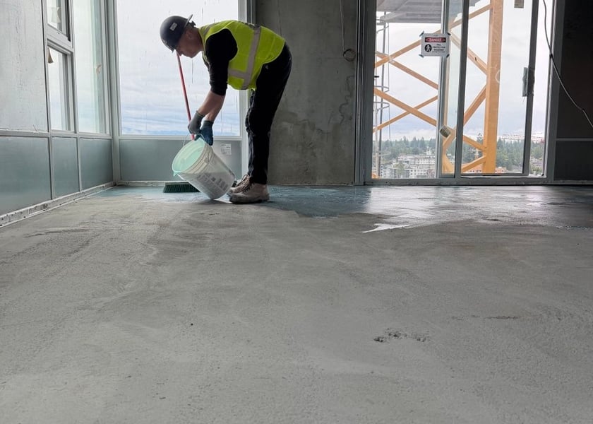 Worker in a hard hat carrying a pail of primer across a bare concrete floor toward a glass door
