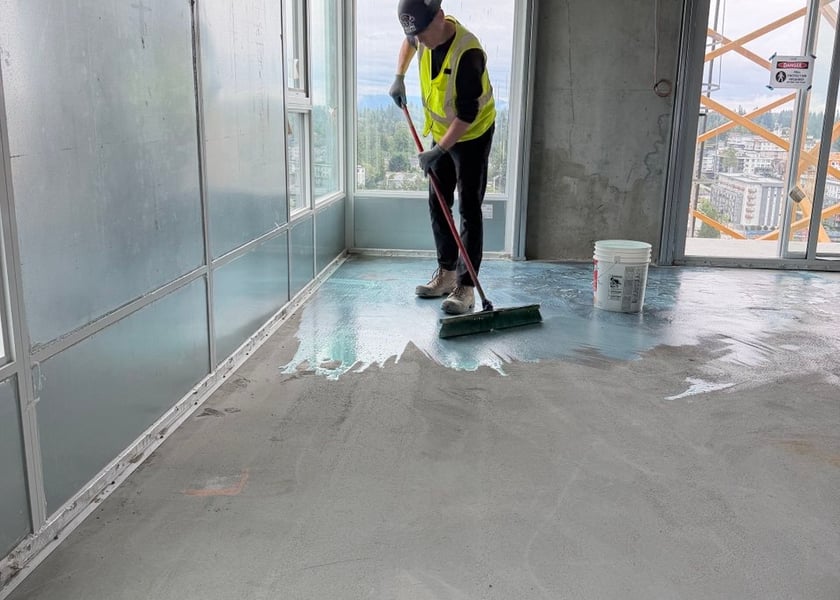 Worker pushing a broom to spread primer across a concrete floor beside a pail, a tower crane in the background