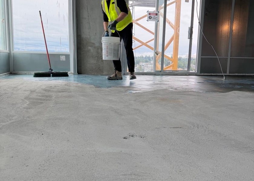 Worker carrying a pail of primer across a bare concrete floor toward a balcony door